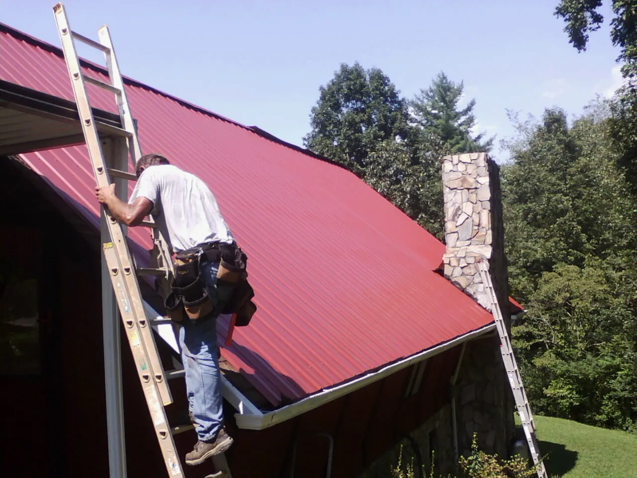Roofer on a ladder beside a red metal roof and stone chimney — Soffit & Fascia Repair by Roofing Brothers in Lake Powell